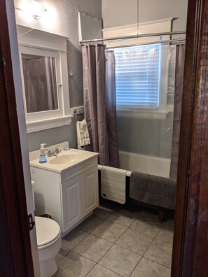 Een badkamer met toilet, wastafel, spiegel, douchegordijn en een raam met jaloezieën.
