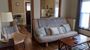 Ein Wohnzimmer mit Sofa, Sessel, Couchtisch und Esstisch. An der Wand hängt eine Uhr, ein Lampenschirm und es gibt ein Fenster mit Vorhängen.