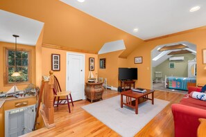 A spacious living room with wooden floors, a television, and a cozy seating area. - Perfectly Content: Walk to Schoolhouse Pond (Chatham)