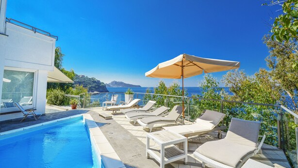 Zona de piscina con sillones y sombrillas, con vistas a un paisaje costero.