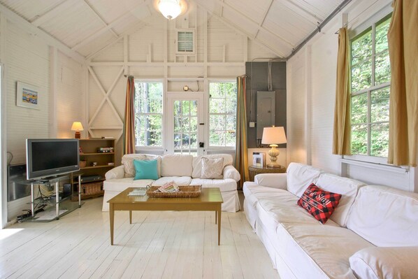 A spacious living room with white furniture, a television, and large windows with curtains.