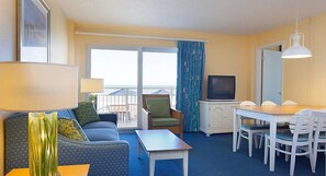 Habitación de hotel con sofá, sillón, mesa de comedor, sillas, televisor y balcón con cortinas.