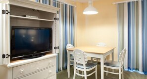 Una habitación de hotel con televisión de pantalla plana, una mesa de madera, sillas blancas y cortinas a rayas.