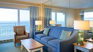 Habitación de hotel con sofá, sillón y dos mesitas auxiliares, ventanas amplias con cortinas y vistas al mar.