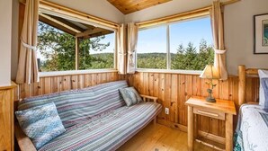 A cozy room with a wooden ceiling and walls, a striped sofa with cushions, a small bedside table with a lamp, and large windows offering a view of trees. - Deer Harbor Cottage on the Bluff (Eastsound)