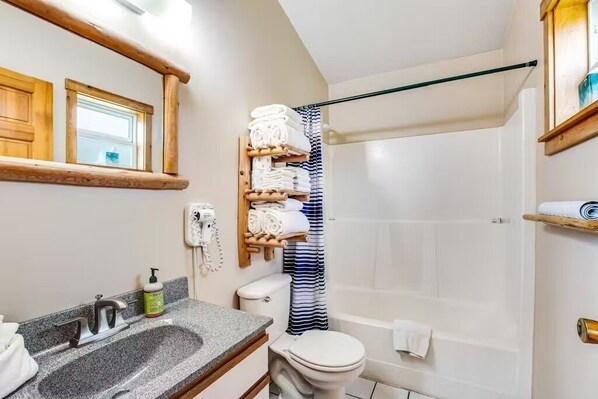 Une salle de bain avec un lavabo en granit, des toilettes, une douche avec un rideau à rayures et une étagère en bois où sont posées des serviettes.