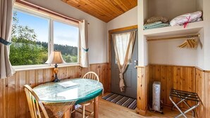 A cozy room with a wooden ceiling, a glass table, chairs, a lamp, a door with curtains, a radiator, and a shelf with folded towels. - Deer Harbor Cottage on the Bluff (Eastsound)
