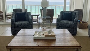 A living room with two patterned armchairs, a wooden coffee table, and a view of the ocean through large windows.
