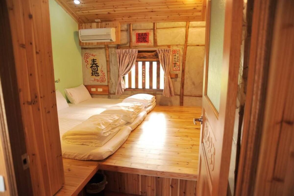 A small, cozy room with a wooden bed, a window with curtains, and a wall-mounted air conditioner.