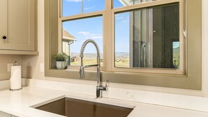 A kitchen with a double sink, a window with a view of a house and landscape, and a potted plant on the windowsill. - *NEW w/ VIEWS* | Modern Mtn. Townhome minutes from RMNP, WP, & Granby Ranch! (Granby)