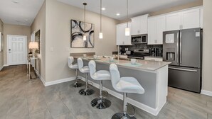 A modern kitchen with white bar stools, stainless steel appliances, and pendant lighting.