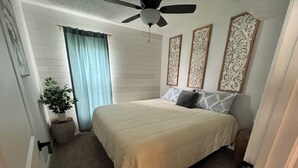 Une chambre à coucher avec un lit, un ventilateur de plafond, des panneaux décoratifs au mur, une plante en pot et une fenêtre avec des rideaux.