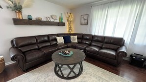 Un salon comprenant un canapé d’angle en cuir marron foncé, une table basse ronde agrémentée d’un élément décoratif central, un tapis à motifs et une étagère murale ornée de divers objets décoratifs.