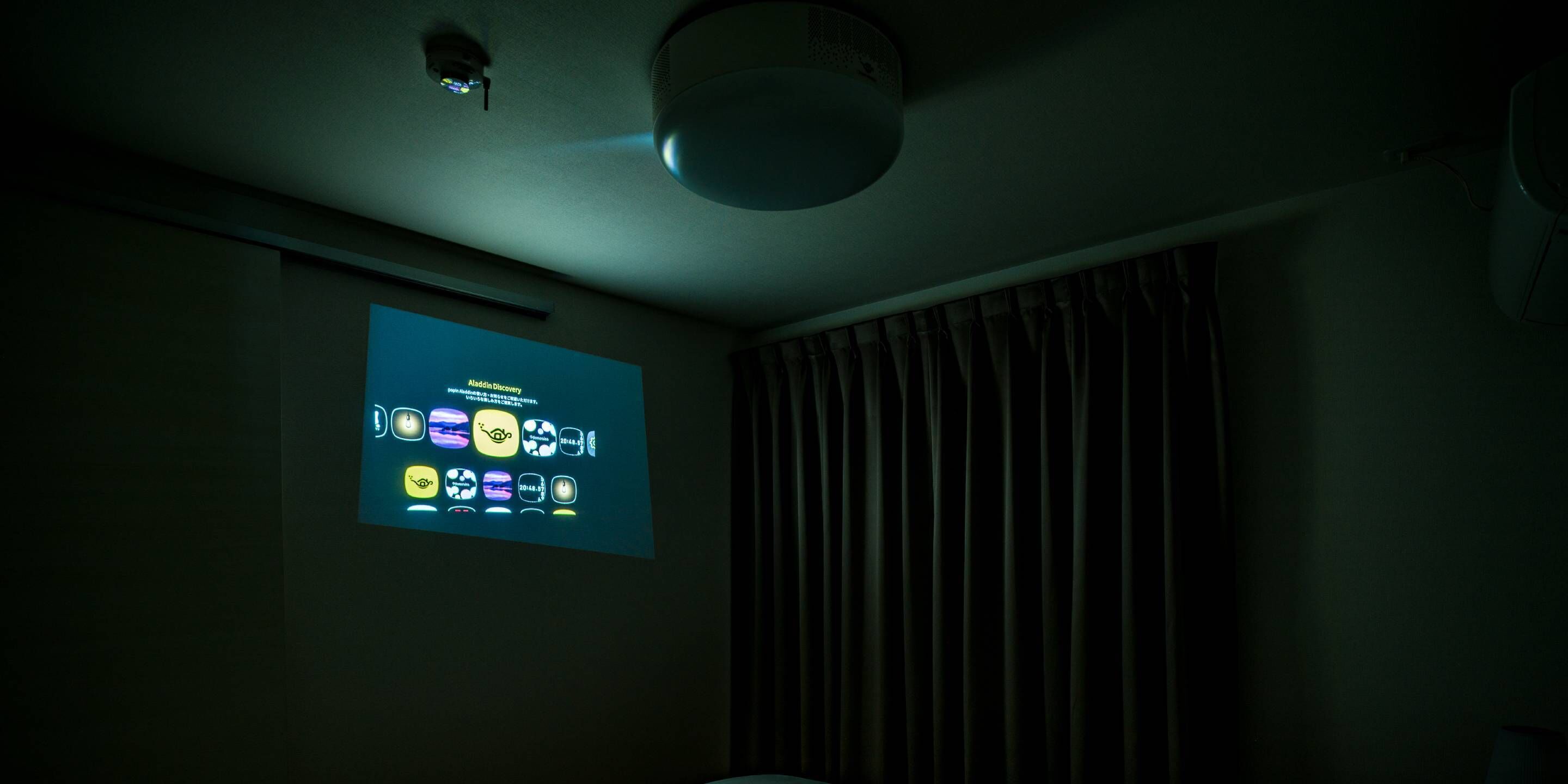 Une pièce sombre avec un écran de projection affichant des icônes, un projecteur fixé au plafond et un rideau.