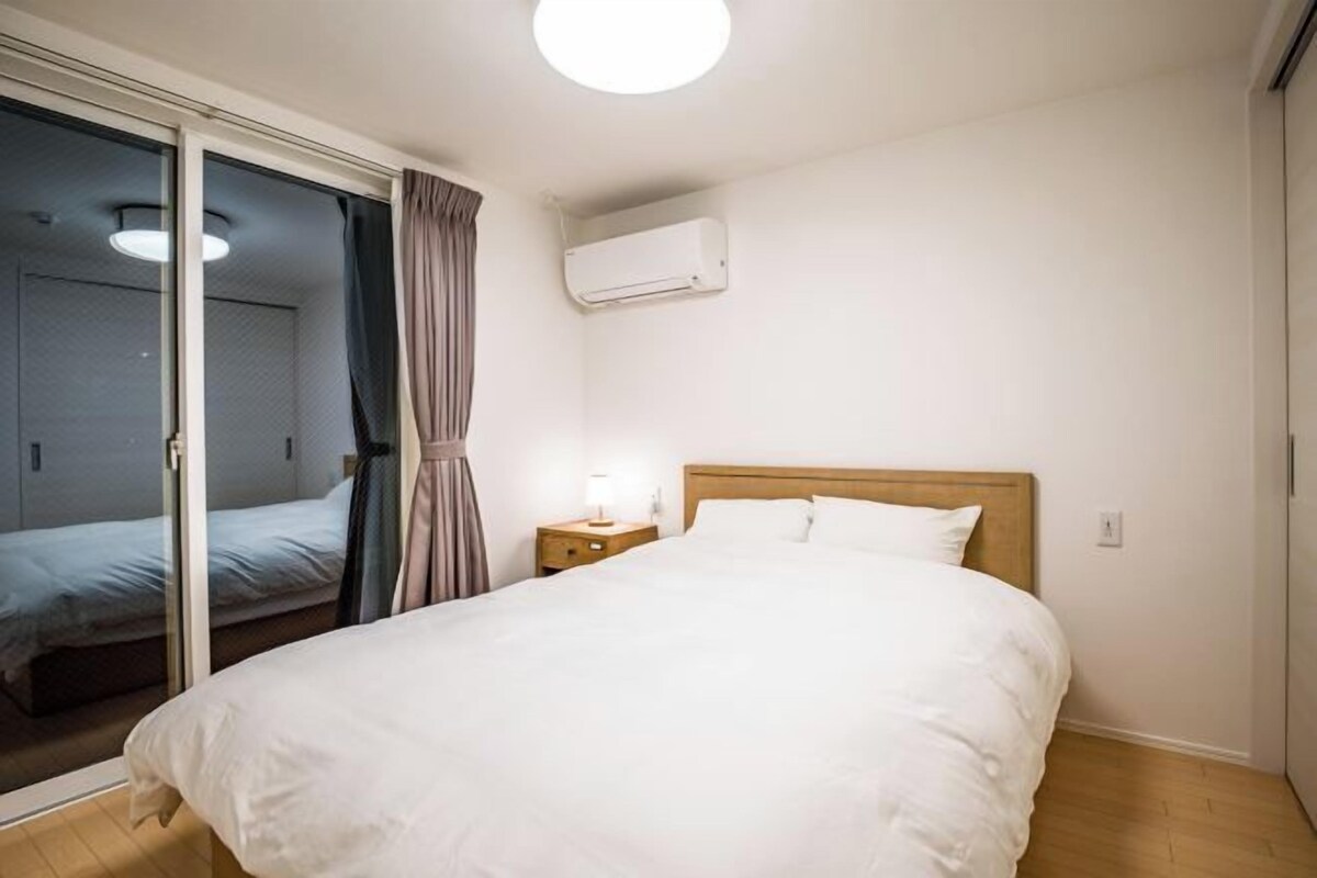 Habitación de hotel con una cama grande, una mesita de noche con lámpara, un equipo de aire acondicionado en la pared y un ventanal corredero que da a otra habitación.