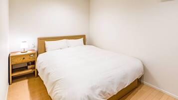 A neatly made bed in a room with a wooden nightstand, a lamp, and a plain white wall.
