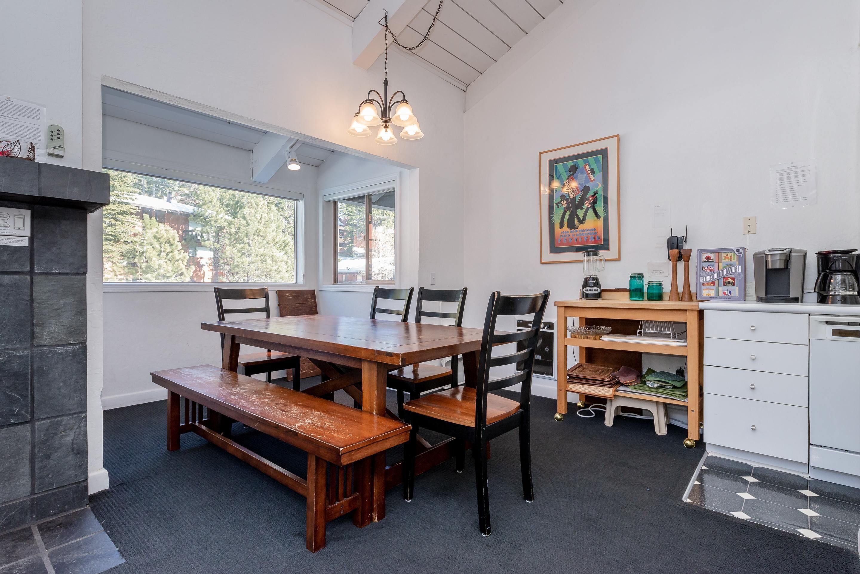 A dining area with a wooden table and benches, a kitchen with white cabinets, a small shelf with kitchen items, and a window with a view of trees.