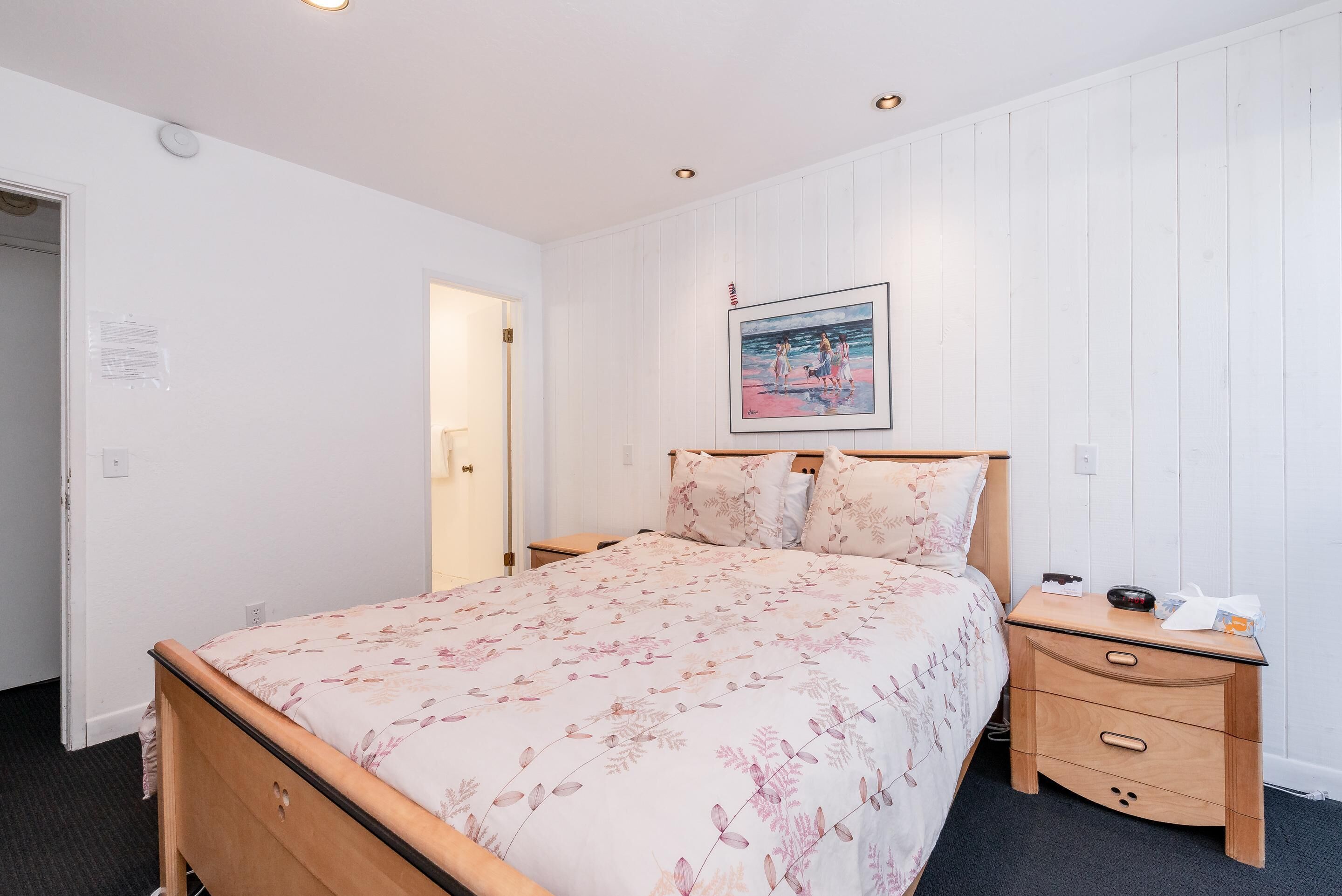 A bedroom with a wooden bed, a nightstand, a framed picture on the wall, and a door to the left.