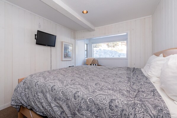 A bedroom with a large bed, a mounted TV, a window with a view of the outdoors, and a wooden ceiling. - Chamonix #62 Bldg. D (Mammoth Lakes)