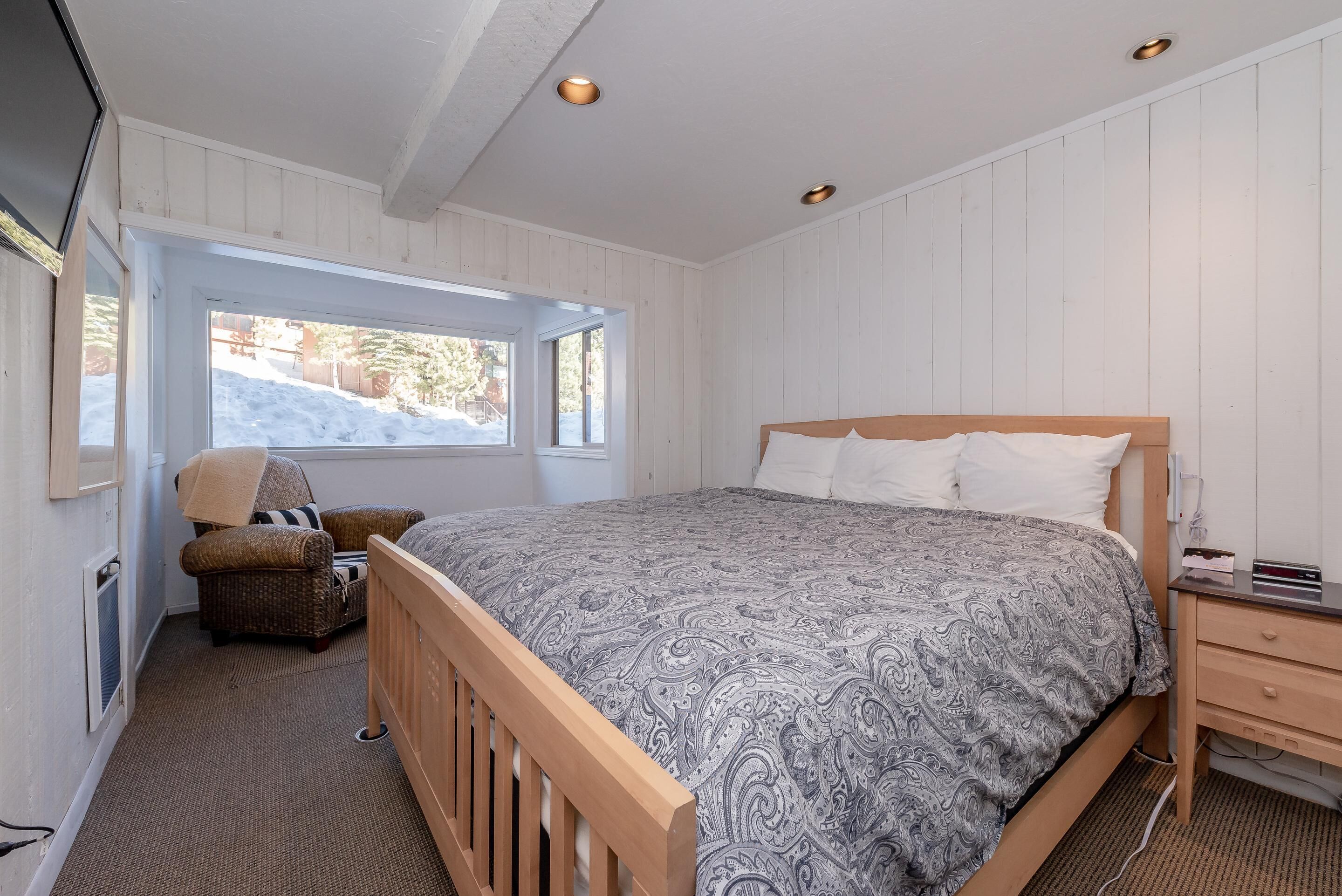 A bedroom with a wooden bed, a nightstand, a chair, and a window with a view of a snowy landscape.