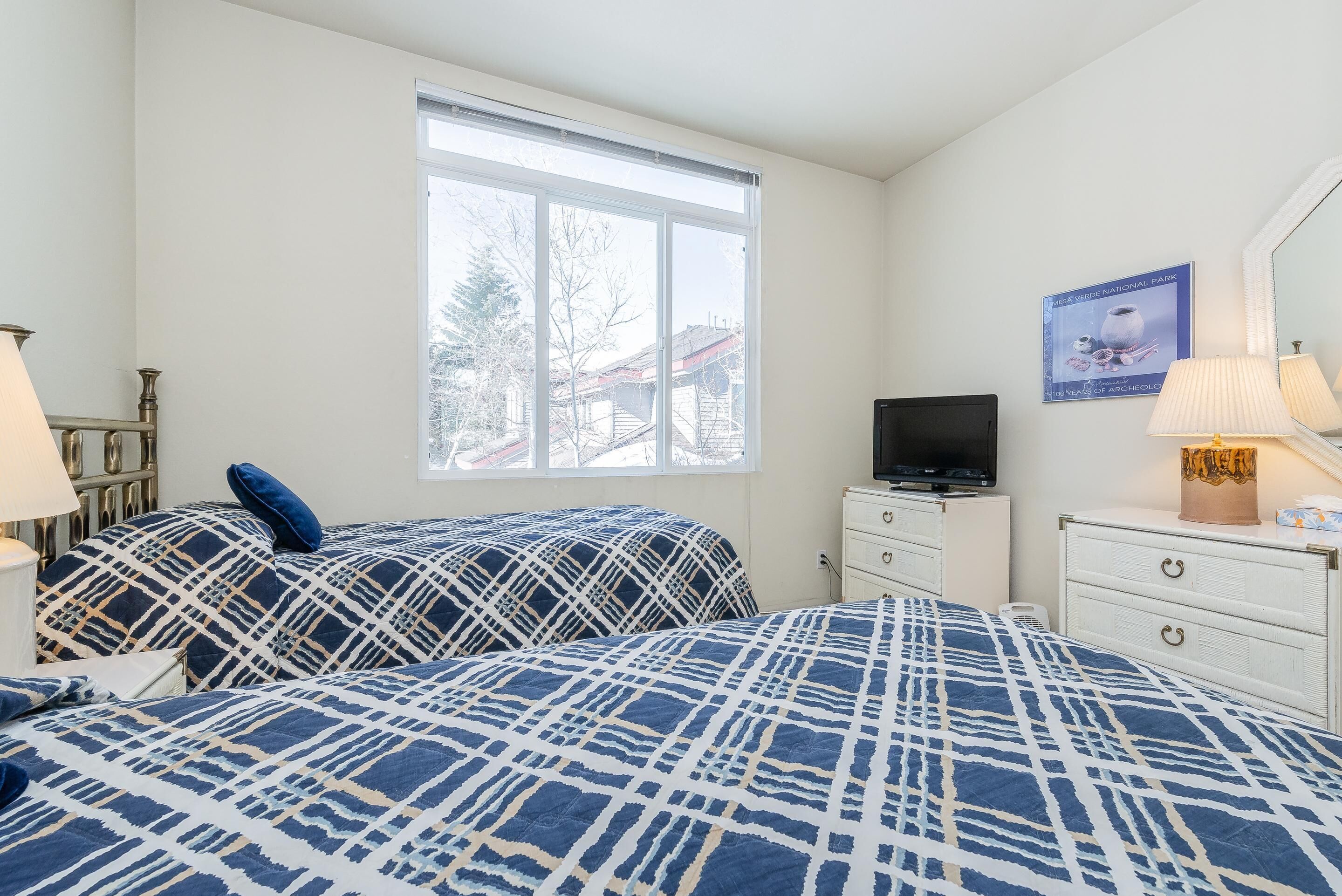 Ein Schlafzimmer mit zwei Betten, einem Fernseher, einem Schminktisch und einem Fenster mit Blick auf Häuser.