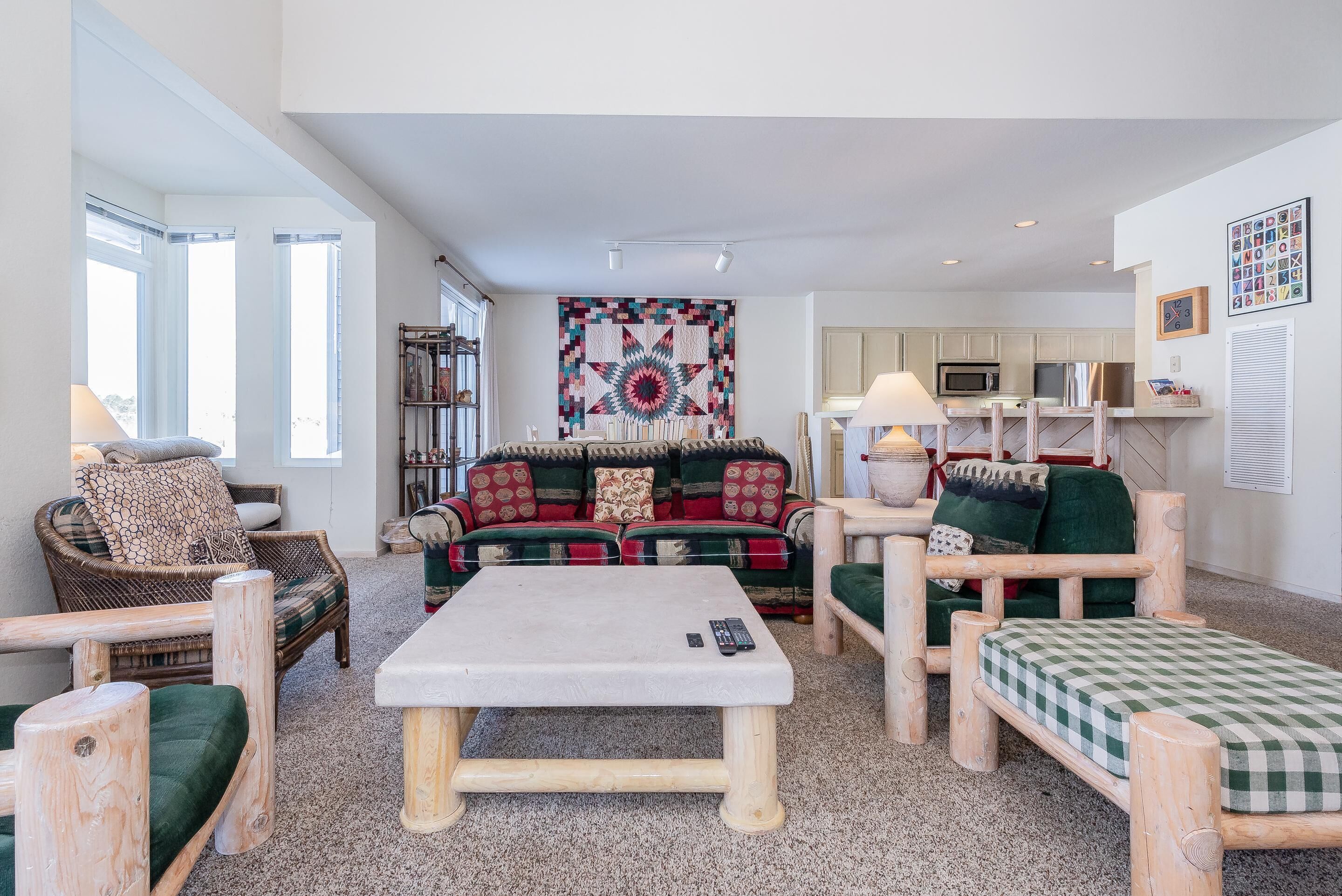 A cozy living room with a large sofa, a coffee table, and wooden furniture. There is a kitchen area in the background with cabinets and a microwave.