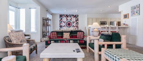 A cozy living room with a large sofa, a coffee table, and wooden furniture. There is a kitchen area in the background with cabinets and a microwave.