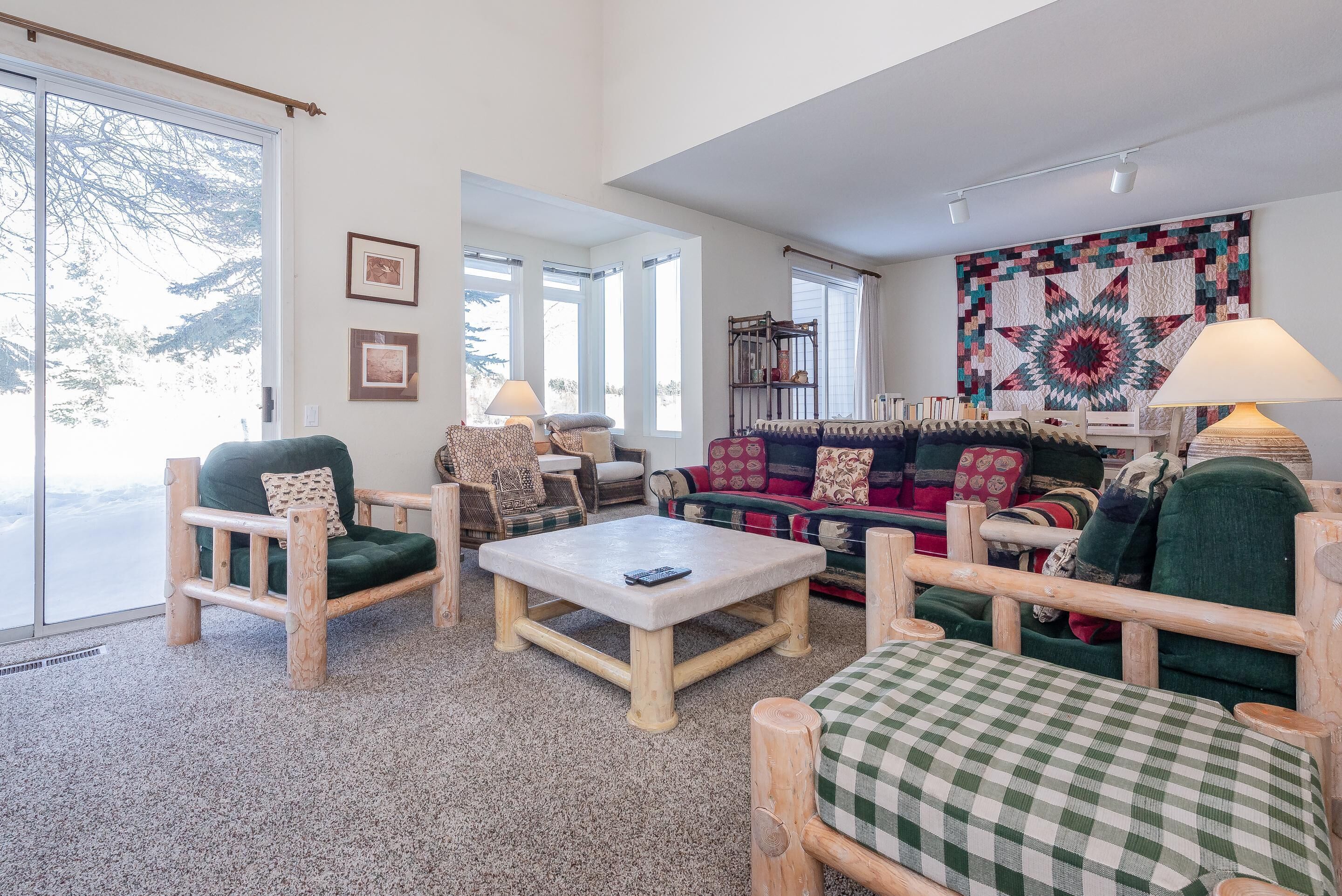 A cozy living room with a large window, a wooden coffee table, and a colorful quilt on the wall.
