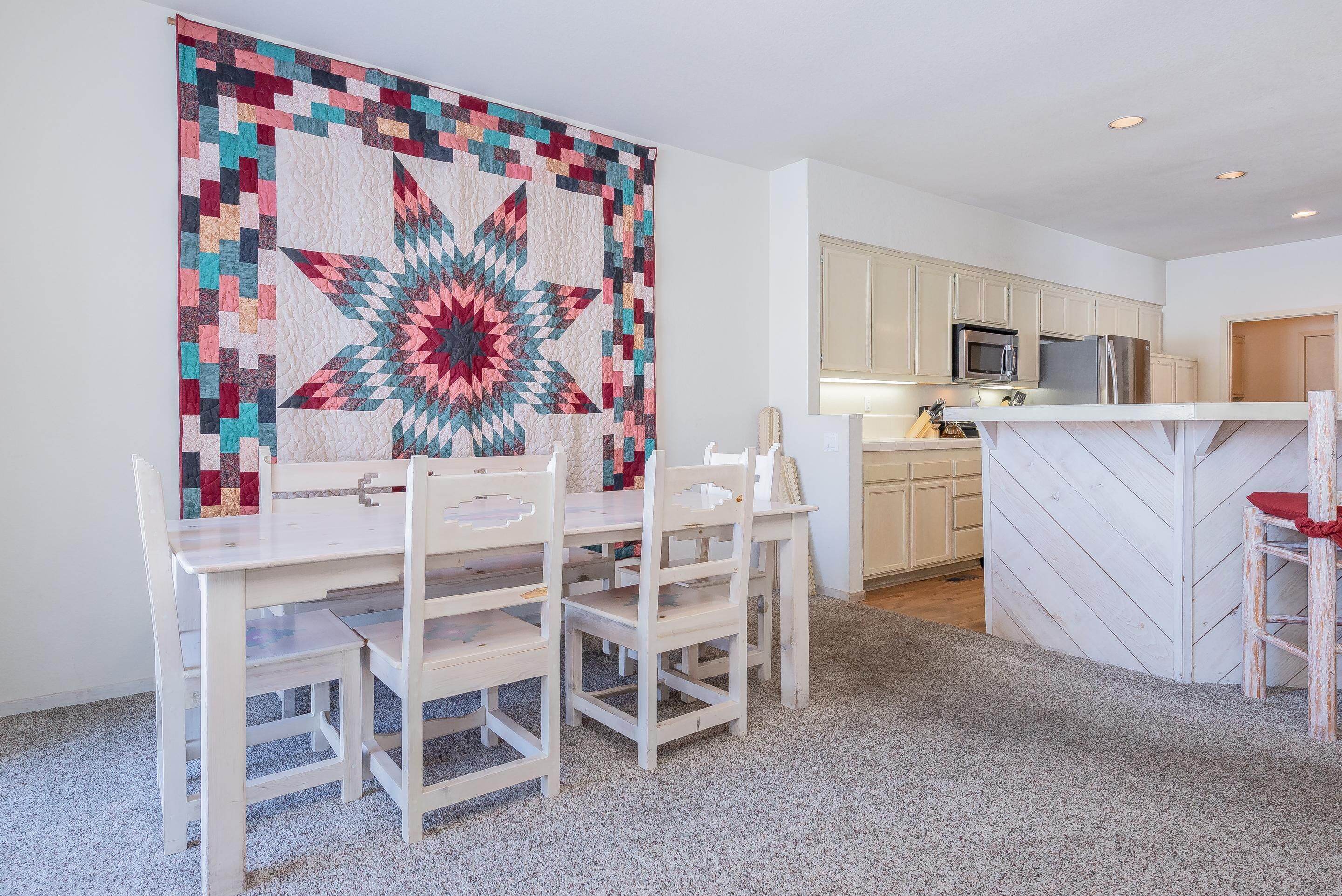 A dining area with a white table and chairs, a large colorful quilt on the wall, and a kitchen in the background.