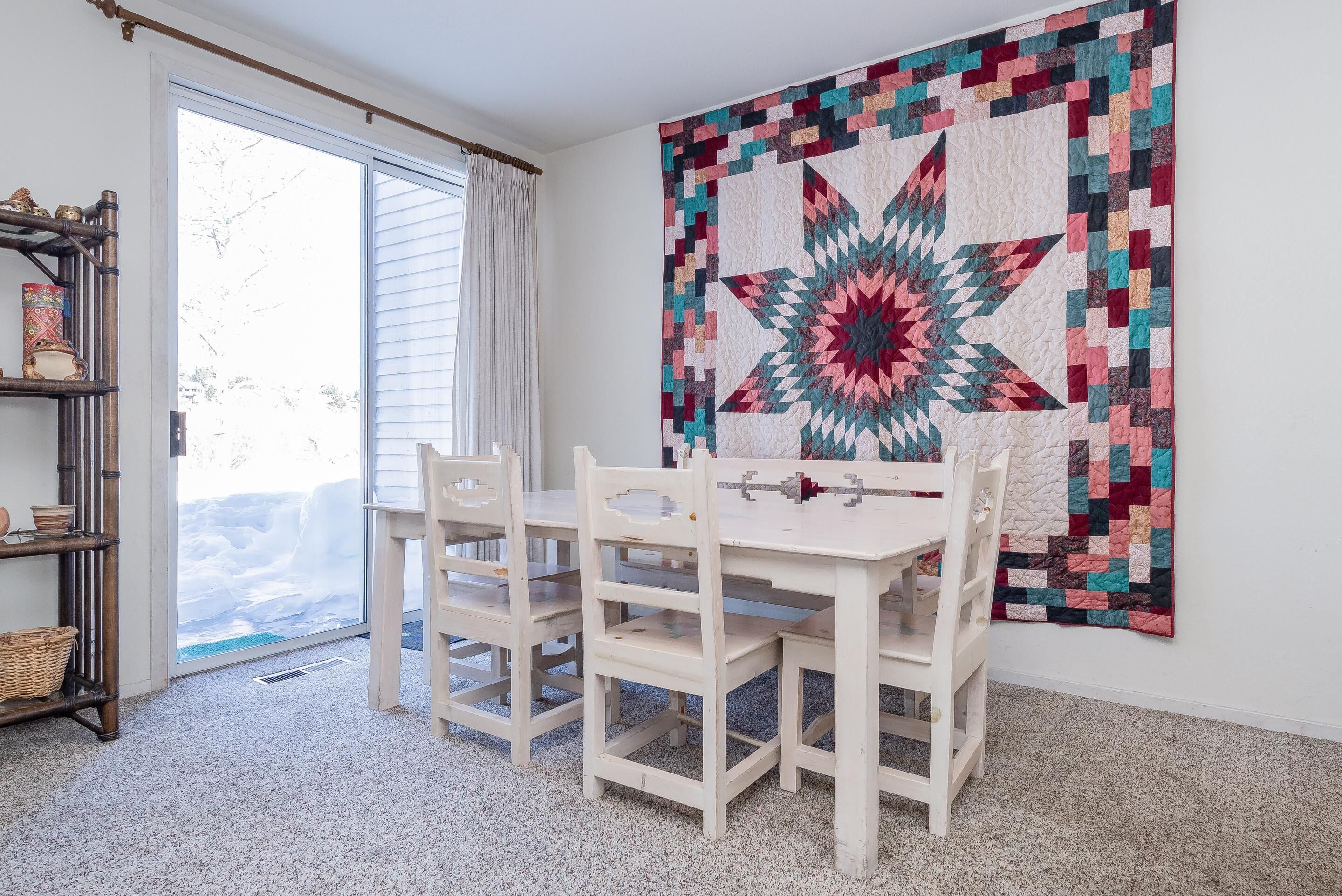 A dining area with a white table and chairs, a large quilt on the wall, and a view through a glass door.