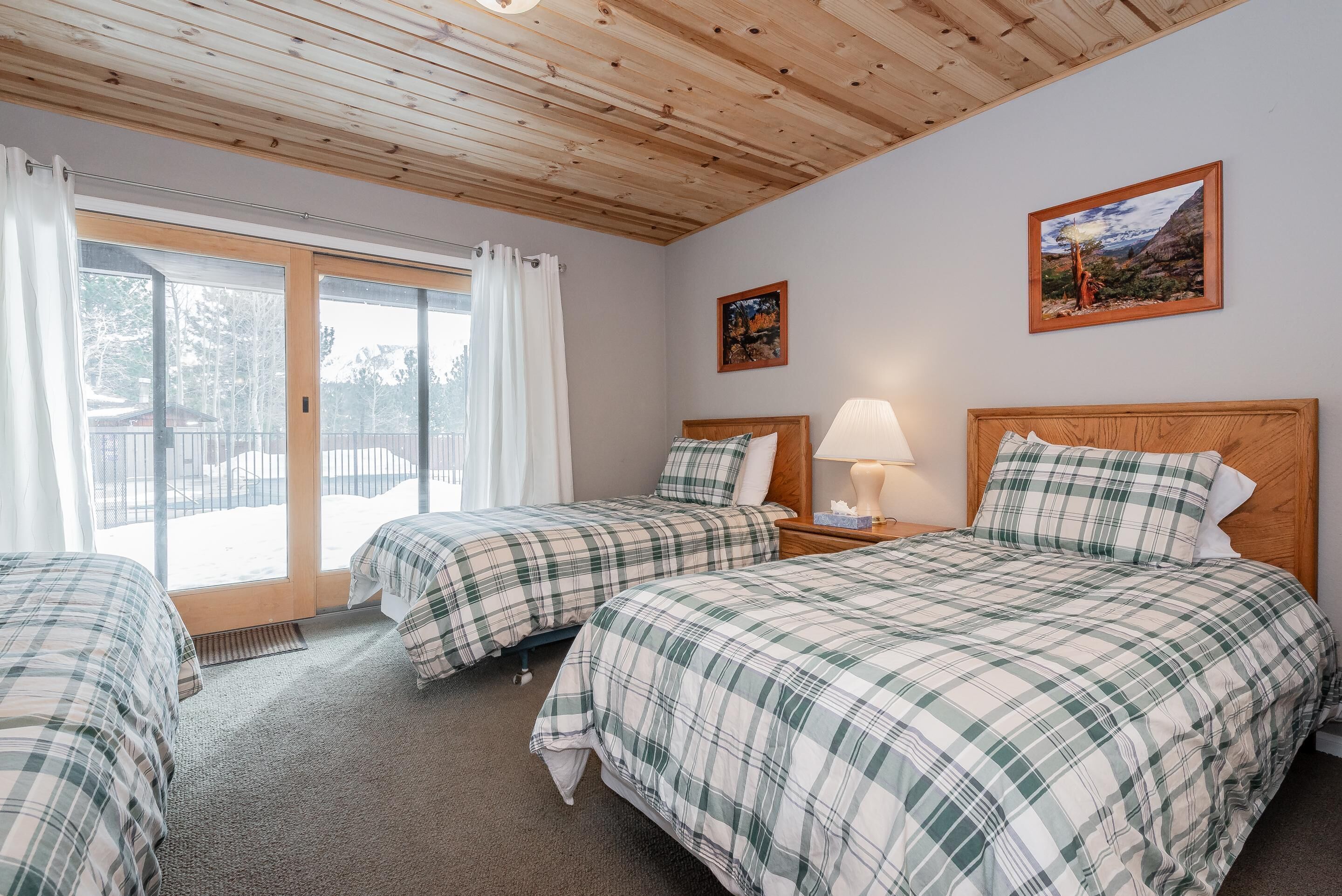 A room with two beds, a wooden ceiling, a large window with curtains, and a framed picture on the wall.