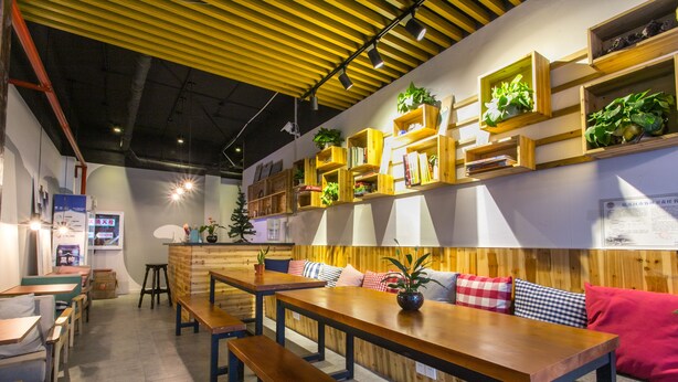 A cozy cafe with wooden tables and benches, shelves with plants, and a yellow ceiling. - Suzhou Blue Gate Hostel (Suzhou)