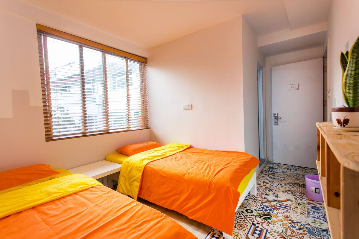 A room with two beds, a window with blinds, a door, and a plant on a wooden dresser.