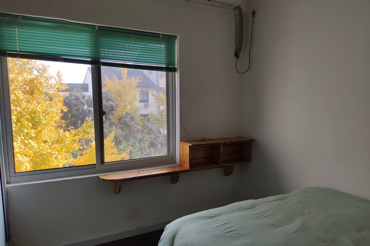 A bedroom with a window, a bed, a wooden shelf, and a wall-mounted telephone.