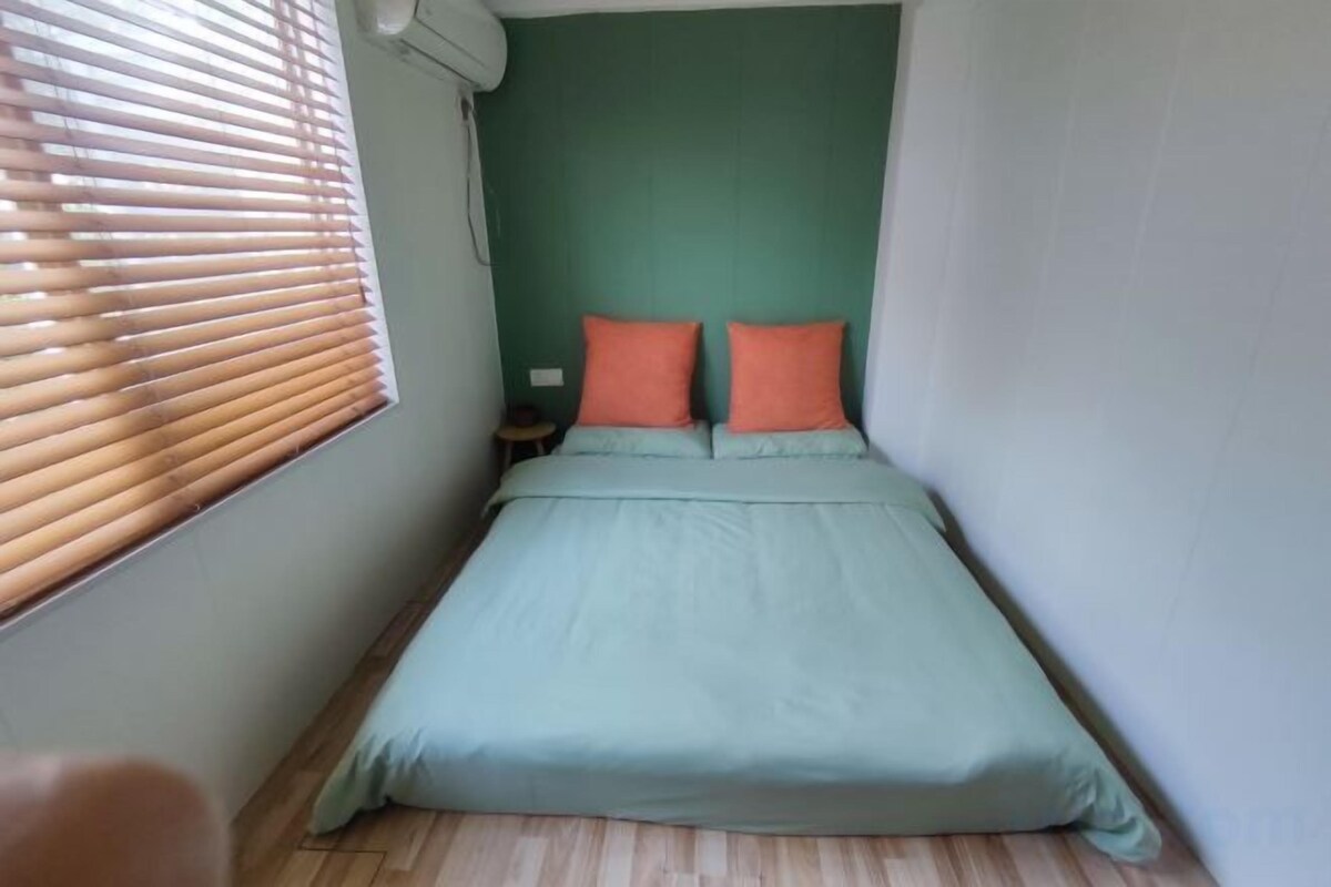 A small, narrow room with a single bed, a nightstand, and a window with blinds.