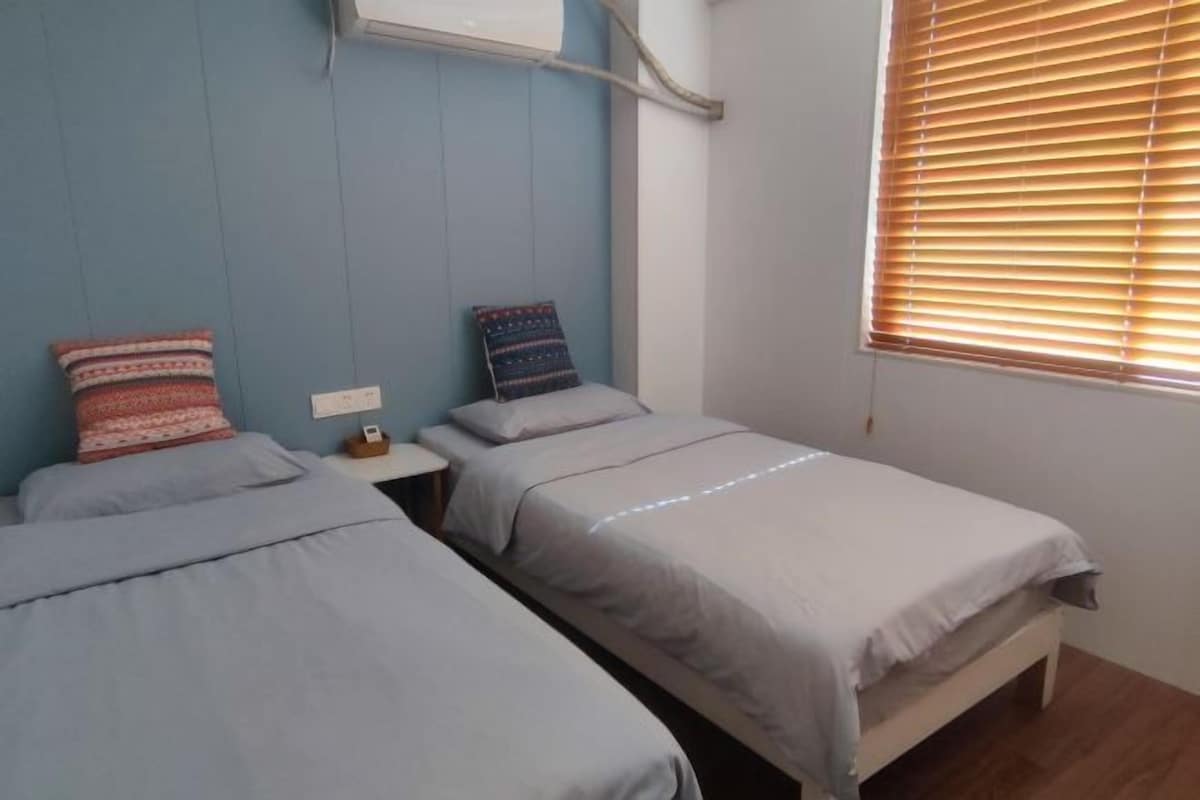 A room with two single beds, a small bedside table with a lamp, and a window with blinds.