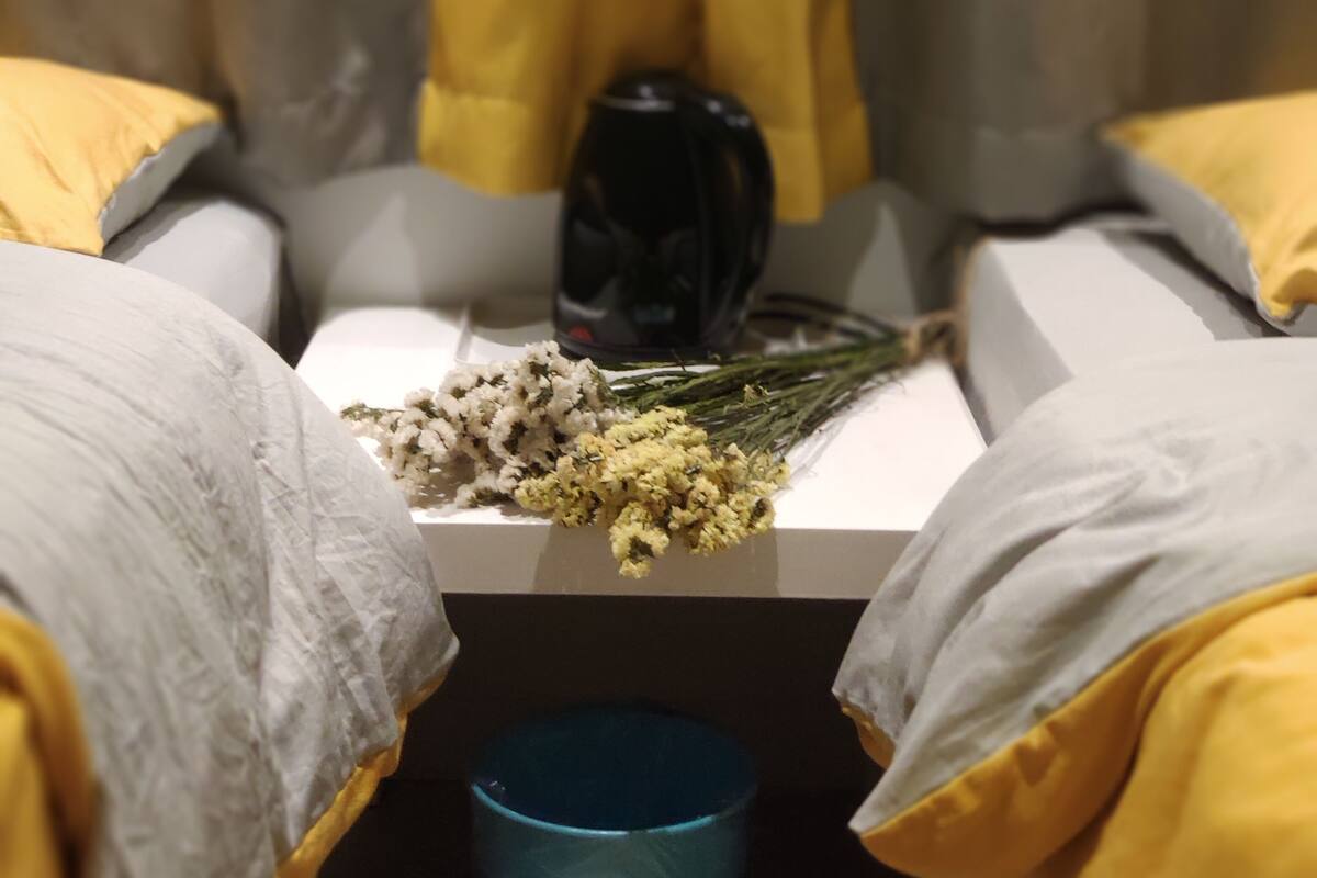 A bed with a white mattress, a blue trash bin, and a black object on the nightstand.