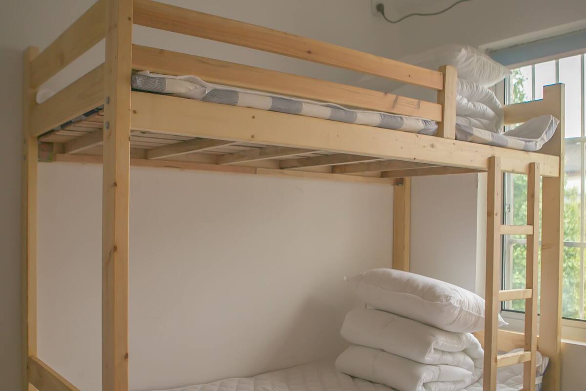A bunk bed with a mattress and pillows, a window with a view, and a trash bin.