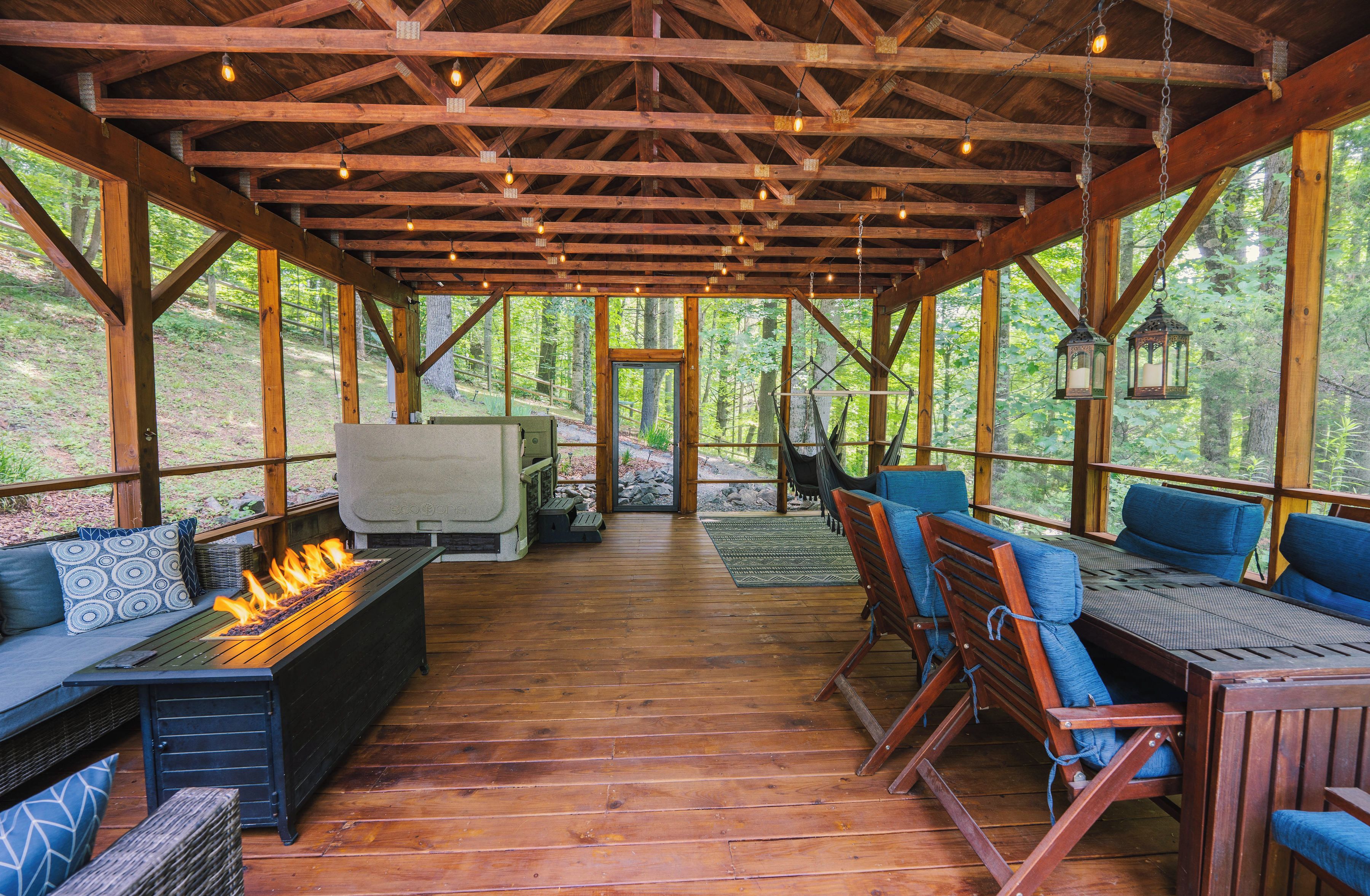 A covered outdoor area with wooden beams, seating, a fire feature, and a view of the forest.
