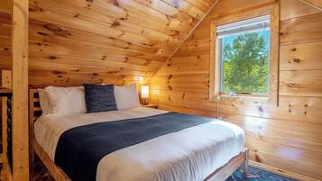 Un dormitorio de cabaña de madera con una cama, almohadas, una mesita de noche, una lámpara y una ventana.