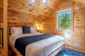 A wooden cabin bedroom with a bed, pillows, a nightstand, a lamp, and a window. - Hot Tub, Views, Romantic Outdoor Lounge Area (Todd)