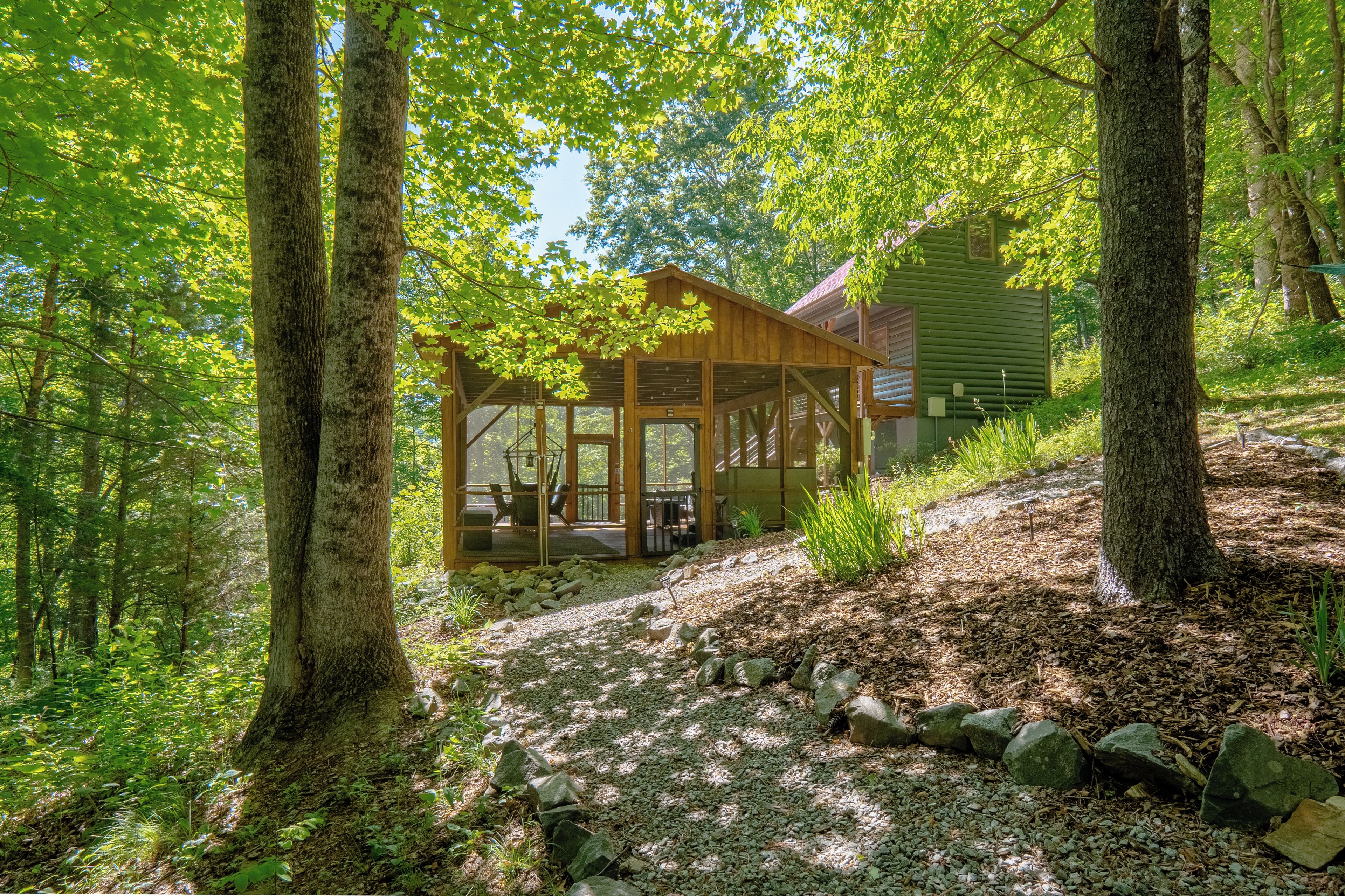 A wooden cabin with a porch, surrounded by trees and a gravel path.