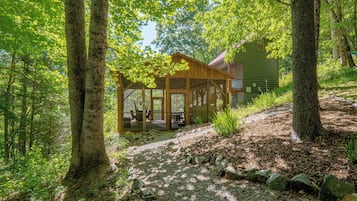 A wooden cabin with a porch, surrounded by trees and a gravel path.