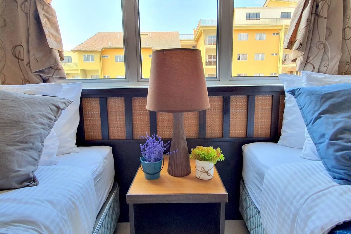 Ein Schlafzimmer mit zwei Betten, einem Nachttisch mit Lampe und Blick auf gelbe Gebäude.