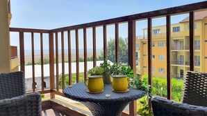En balkon med fletstole, et rundt bord med gule krukker og udsigt til en strand og boliger.