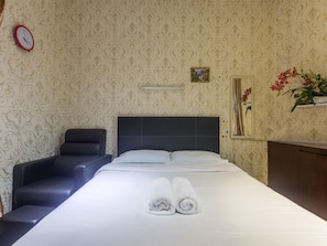Una habitación de hotel con una cama grande, cabecero, mesita de noche, una silla, un reloj, un cuadro enmarco y una planta en maceta.