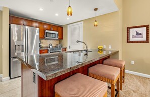 A modern kitchen with a stainless steel refrigerator, microwave, and stove, a large island with bar stools, and pendant lighting. - Maui Resort Rentals:  Honua Kai Hokulani 111 – Ground Floor Interior Courtyard 2BR, Steps to Pools, Beach, and BBQs, Large Lawn Area! (Lahaina)