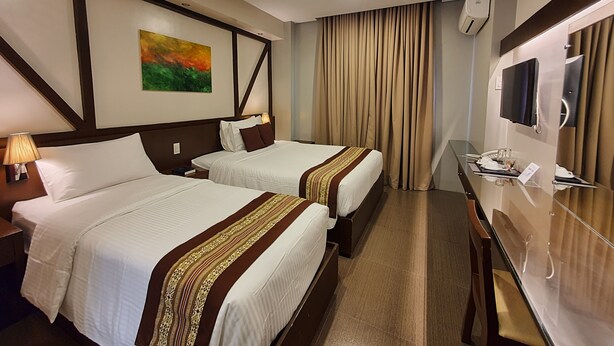 A hotel room with two beds, a desk, a mirror, and a painting on the wall. - Missio Dei Hotel (Dipolog)