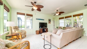 A living room with a sofa, a television, wicker furniture, and ceiling fans. - 🏖️Private Kona Home w/ Pool, Sand Court & Ocean View Lanai | Nakukui Ekolu (Kailua Kona)