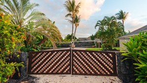 A gated entrance with a wooden lattice fence, surrounded by tropical vegetation and a stone wall. - 🏖️Private Kona Home w/ Pool, Sand Court & Ocean View Lanai | Nakukui Ekolu (Kailua Kona)